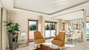 A modern living room with a glass coffee table, two brown armchairs, a glass coffee table, a potted plant, and a view of the city through large windows.