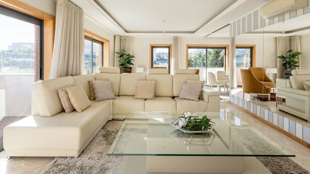 A modern living room with a beige sectional sofa, a glass coffee table, and a view of the city through large windows.