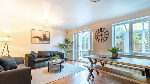 A living room with a sofa, coffee table, and a dining table with benches. There are plants, a clock, and a window with a view. - Lovely 5 Bed House in Egham near Heathrow & Virginia Water (Egham)