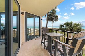 A balcony with a table and chairs, a glass door leading to a pool, and a view of the beach and palm trees.