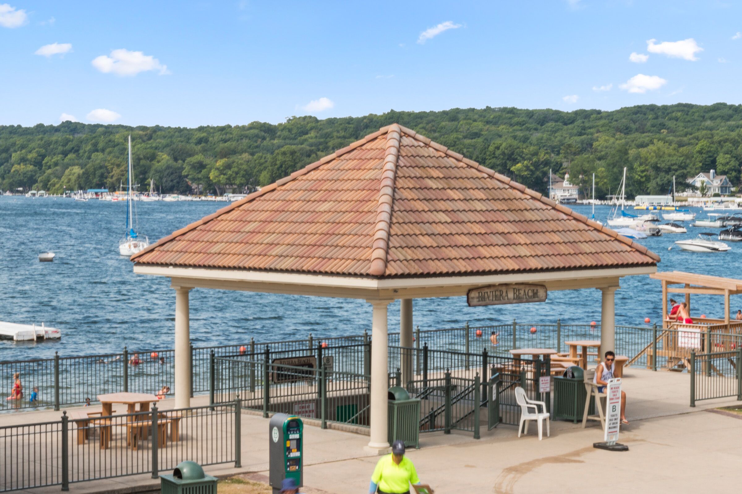 A gazebo with a tiled roof overlooking a marina with boats and a forested hill in the background.