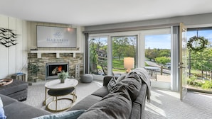 Una sala de estar moderna con chimenea, un sofá, una mesa de centro y vistas a un paisaje verde a través de amplios ventanales correderas.