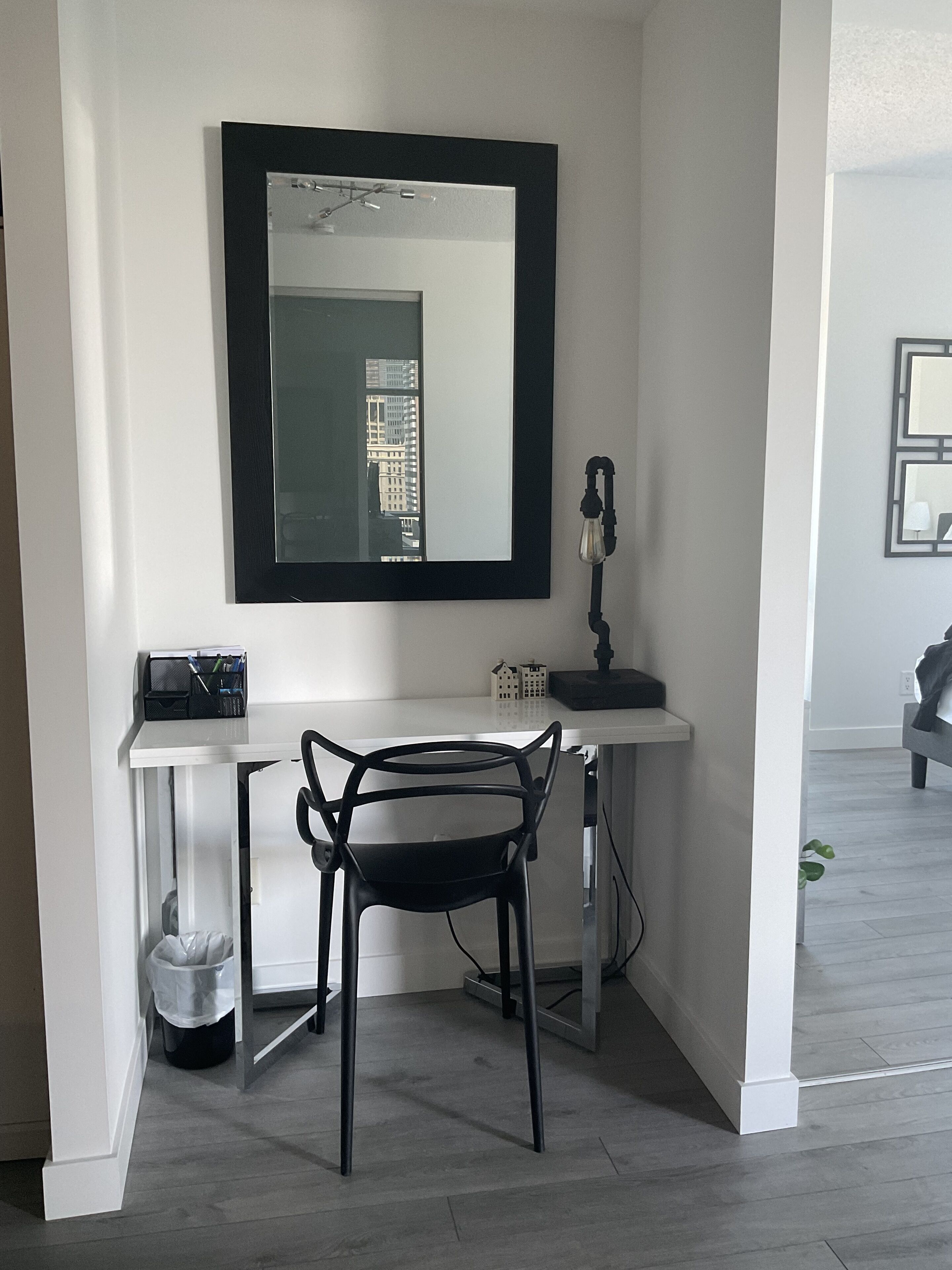A modern, minimalistic room with a white desk, a black chair, a large mirror, and a small lamp.
