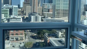 Une vue sur une ville à travers une fenêtre, avec des bâtiments modernes, dont une tour remarquable coiffée d’un sommet rouge et blanc.
