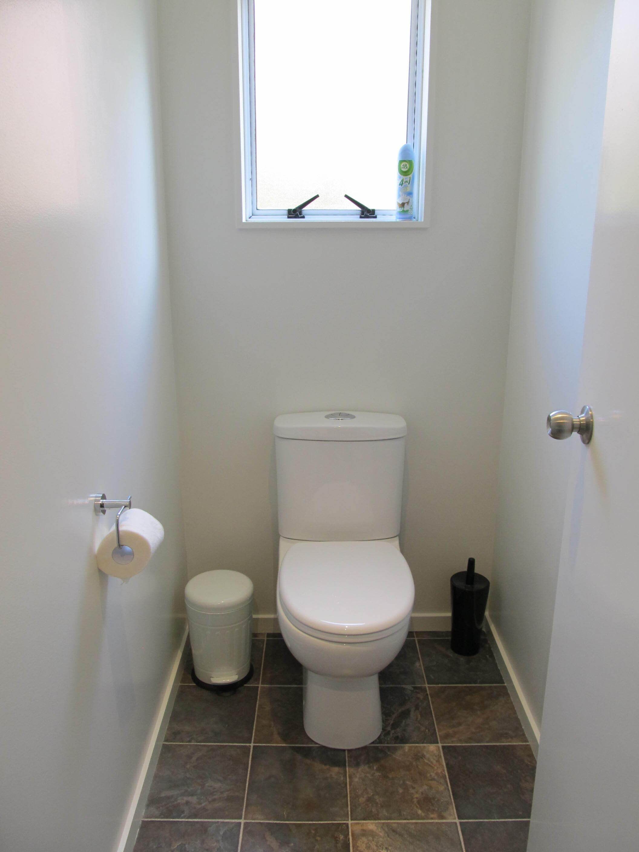 A small, clean bathroom with a toilet, a trash can, and a window above the toilet.