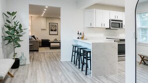 Een moderne keuken met witte kasten, een centraal kookeiland met barstoelen, en een zithoek met een bank en salontafel.