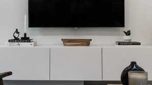 Een moderne woonkamer met een flatscreen televisie die op een wit kastje is bevestigd, een zwarte salontafel met een kaars en een boek, en een houten stoel.