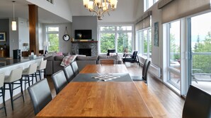 A spacious living room with a large wooden dining table, comfortable seating, a fireplace, and large windows offering a view of the outdoors.