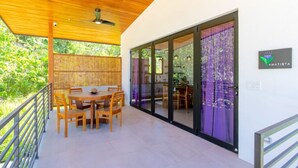 A covered patio area with a wooden table and chairs, a ceiling fan, and a view of the interior through glass doors. - La Joya Amatista - Walk to Beach (Nosara)