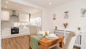 A modern kitchen with white cabinets, a wooden dining table set with plates, glasses, and candles, and a cozy chair. - The Coastal Hideaway (Hornsea)