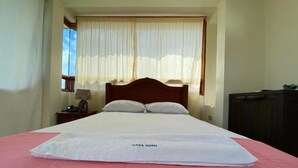 Cama bem arrumada com colcha rosa e branca, cabeceira de madeira e janela com cortinas leves.
