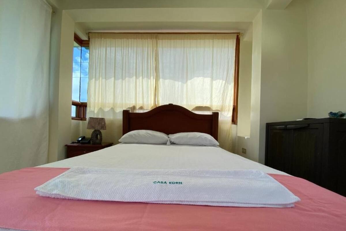Ein ordentlich bezogenes Bett mit einer rosa-weißen Tagesdecke, einem Holz-Kopfteil und einem Fenster mit transparenten Vorhängen.