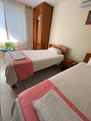 A hotel room with two beds, a wooden wardrobe, a green chair, and a window with curtains. - Hostal Casa Eden Galapagos (Puerto Ayora)