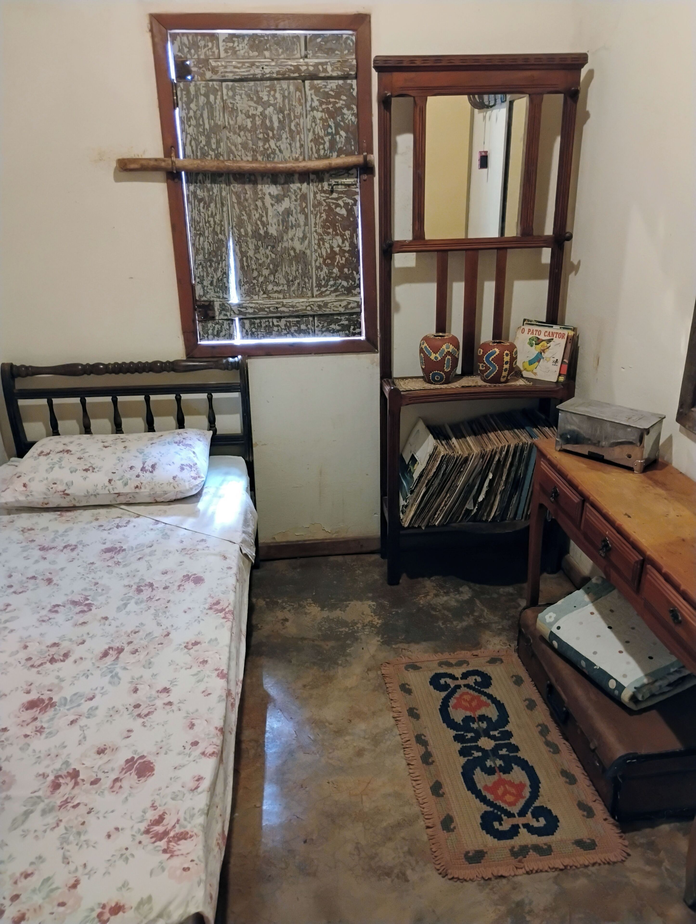 Una camera da letto con un letto, un comò in legno, una mensola con libri e un vaso, e una finestra con tenda.