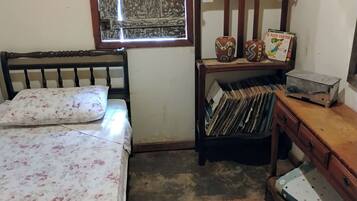 Una camera da letto con un letto, un comò in legno, una mensola con libri e un vaso, e una finestra con tenda.