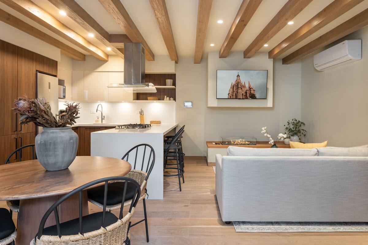 Une cuisine moderne avec des poutres en bois, un coin repas avec une table ronde et des chaises, et un salon avec un canapé et un tableau représentant un bâtiment.