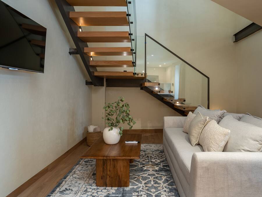 Un salón moderno con una escalera de madera, un televisor de pantalla plana montado en la pared, un sofá color beige y una mesa de centro con un jarrón y un control remoto.