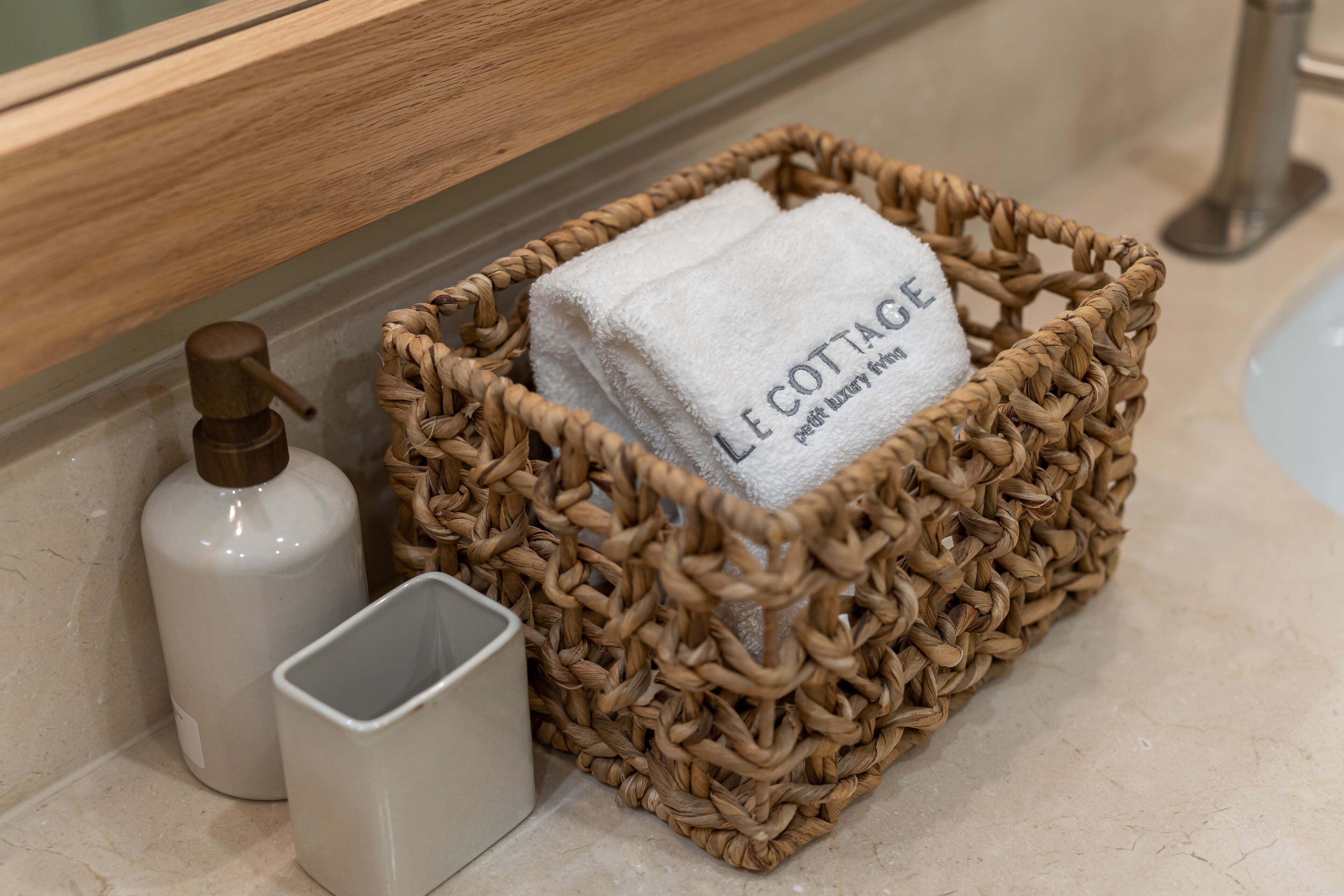 A bathroom with a woven basket containing a white towel labeled "L COTTAGE" and a white soap dispenser.