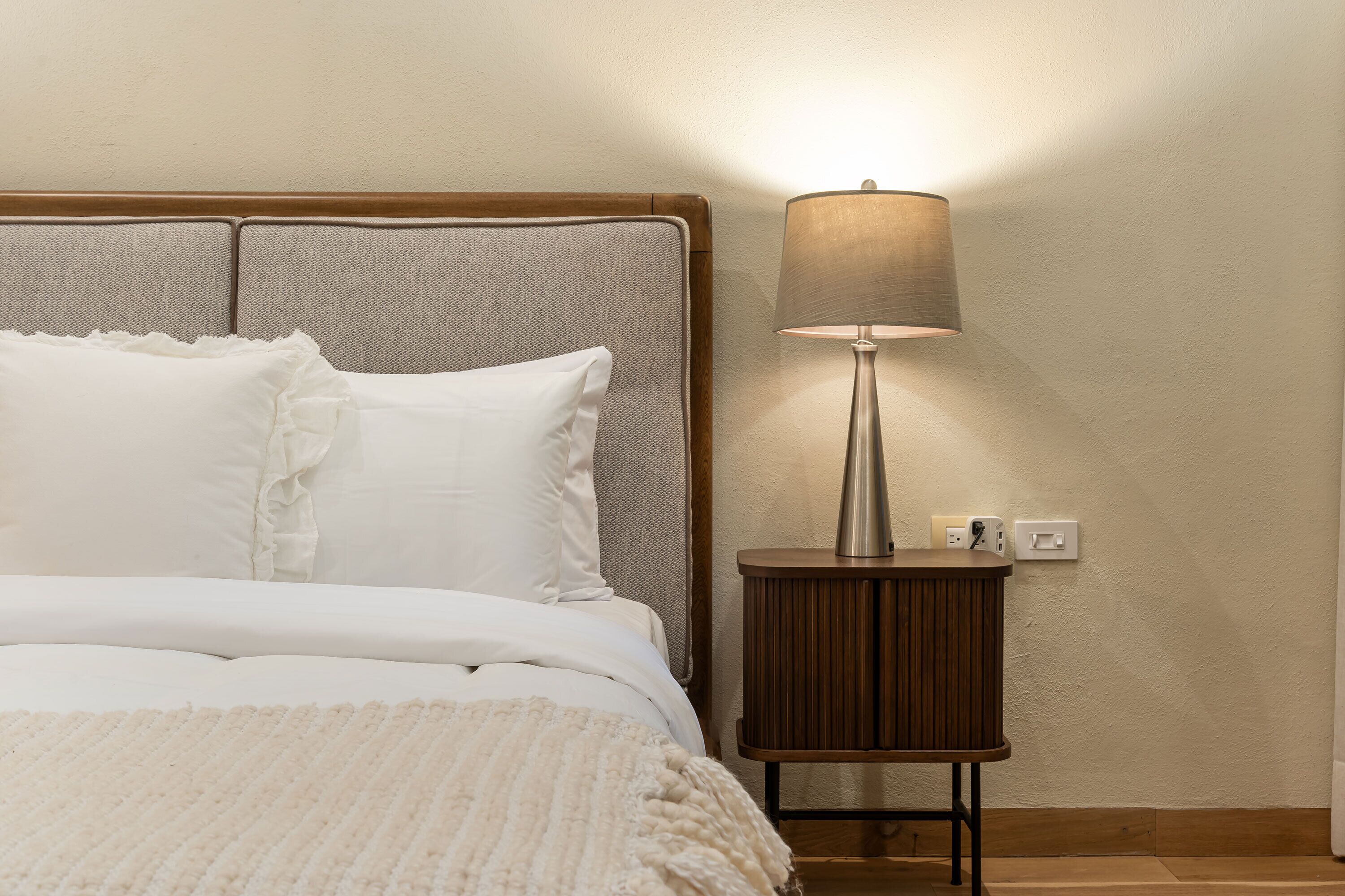 A neatly made bed with white bedding and pillows, a bedside lamp, and a wooden nightstand.