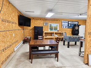 A room with a wooden interior, a black refrigerator, a wooden table with chairs, a foosball table, and a dartboard. - Anglers Lodge 3 (Webster)