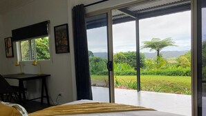 A room with a bed, a desk with two lamps, a window with a curtain, and a view of a green landscape. - NEW -The Sugar Shack, Ocean views over cane fields from your outdoor Spa Bath  (Miallo)