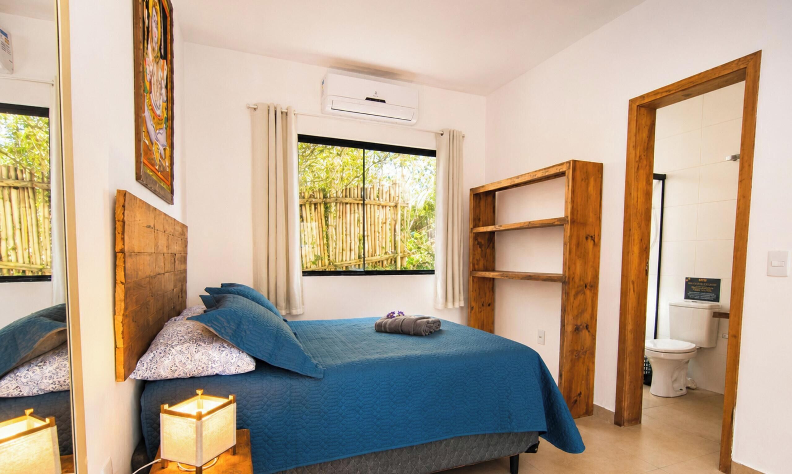 A bedroom with a bed, bedside lamp, window, air conditioning unit, and a view of greenery outside.
