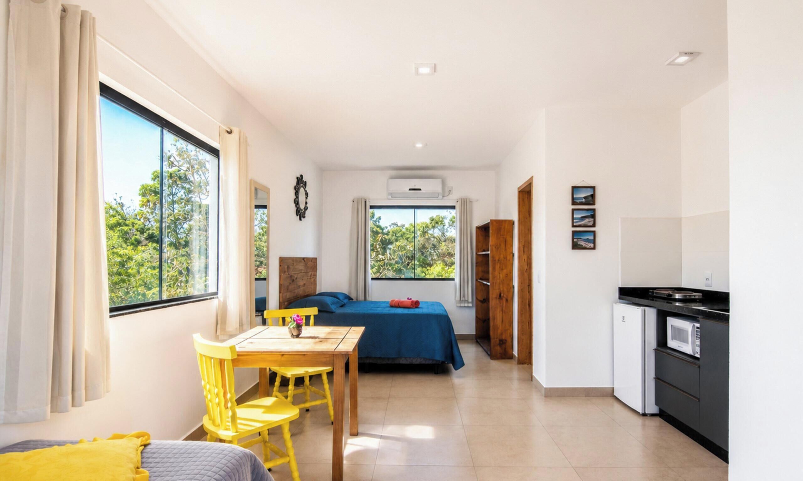 A compact room with a bed, a table with chairs, a kitchenette, and large windows.