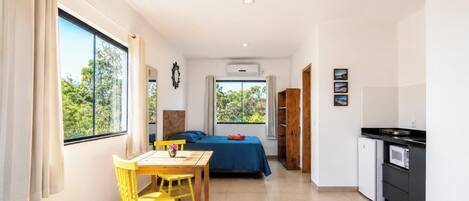 A compact room with a bed, a table with chairs, a kitchenette, and large windows.