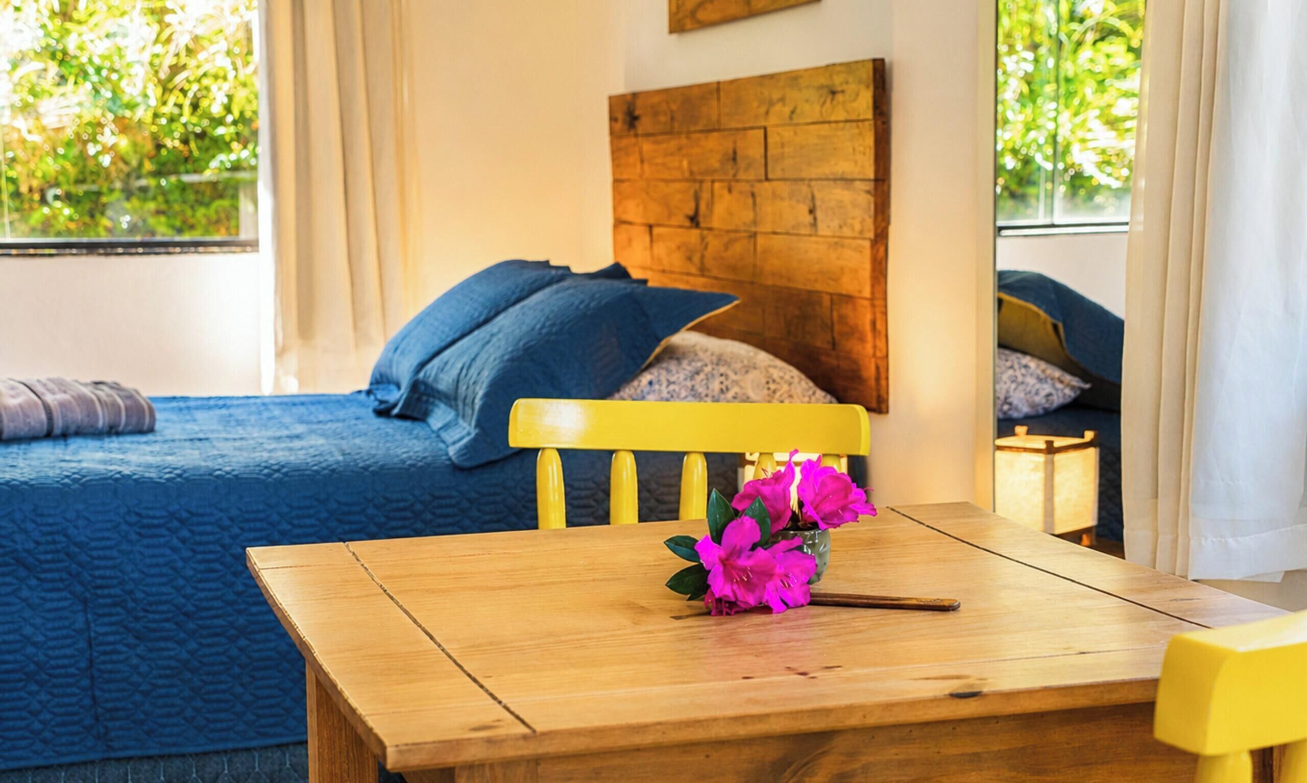Quarto com mesa de madeira, cama com roupa de cama azul e janela com cortinas.