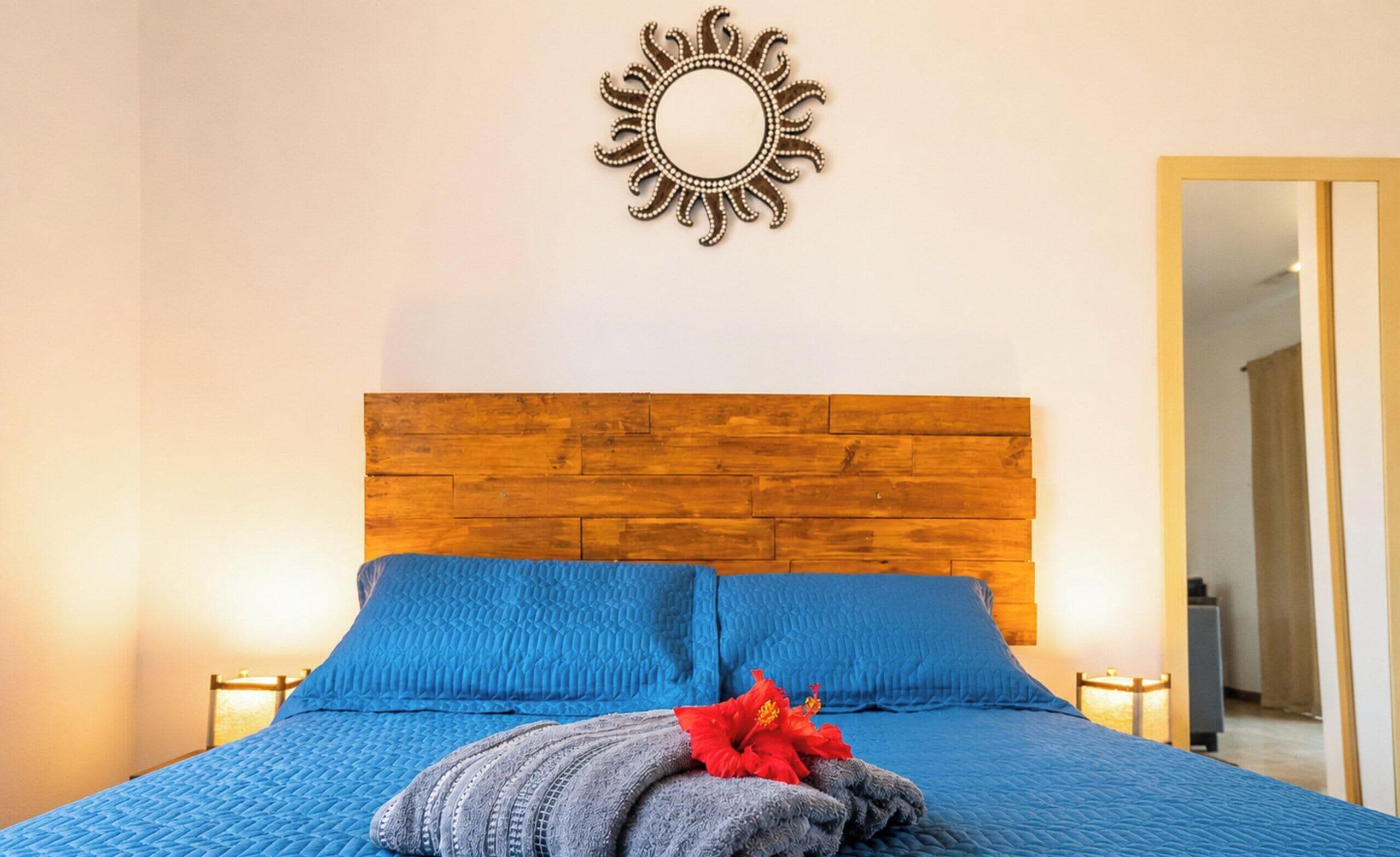 A bedroom with a wooden headboard, a blue bedspread, two pillows, a decorative wall mirror, and bedside lamps.