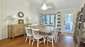 A dining area with a wooden table, white chairs, a ceiling fan, and a view of the outdoors through large windows.