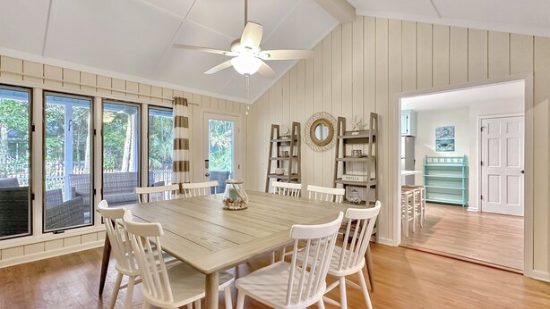 A dining area with a wooden table and white chairs, a ceiling fan, and a view of the outdoors through large windows.
