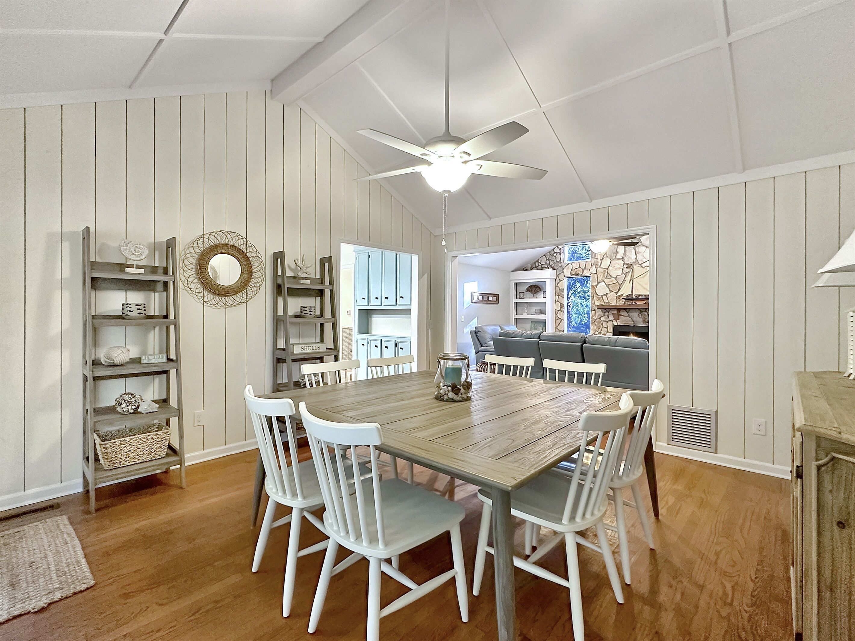 A dining area with a wooden table and white chairs, a ceiling fan, and a kitchen in the background.