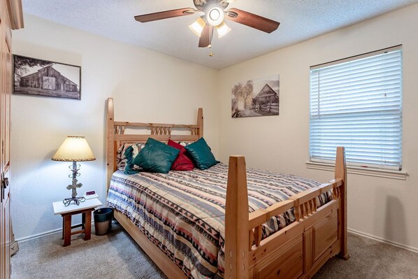 A bedroom with a wooden bed, a ceiling fan, a bedside table with a lamp, and two wall-mounted pictures.
