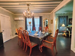 A dining room with a long table set for six, surrounded by wooden chairs. A chandelier hangs from the ceiling, and there is a window with patterned curtains.