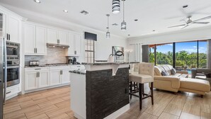 A modern kitchen with white cabinets, a central island with a black backsplash, and a dining area with a beige sofa and a view of the outdoors. - Fairway Evergreen: Theater, Themed Bedroom, Private Pool, Water Park, Near Disney (Kissimmee)
