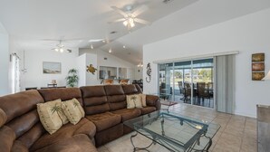 A spacious living room with a large brown sectional sofa, glass coffee table, ceiling fans, and a view to the outdoor patio area. - Freshwater Canal, Heated Pool & Brand New Appliances - Villa Seahorse Sanctuary - Roelens (Cape Coral)