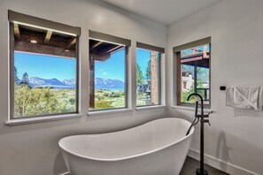 A modern bathroom with a freestanding bathtub, large windows offering mountain views, and a visible outdoor structure.