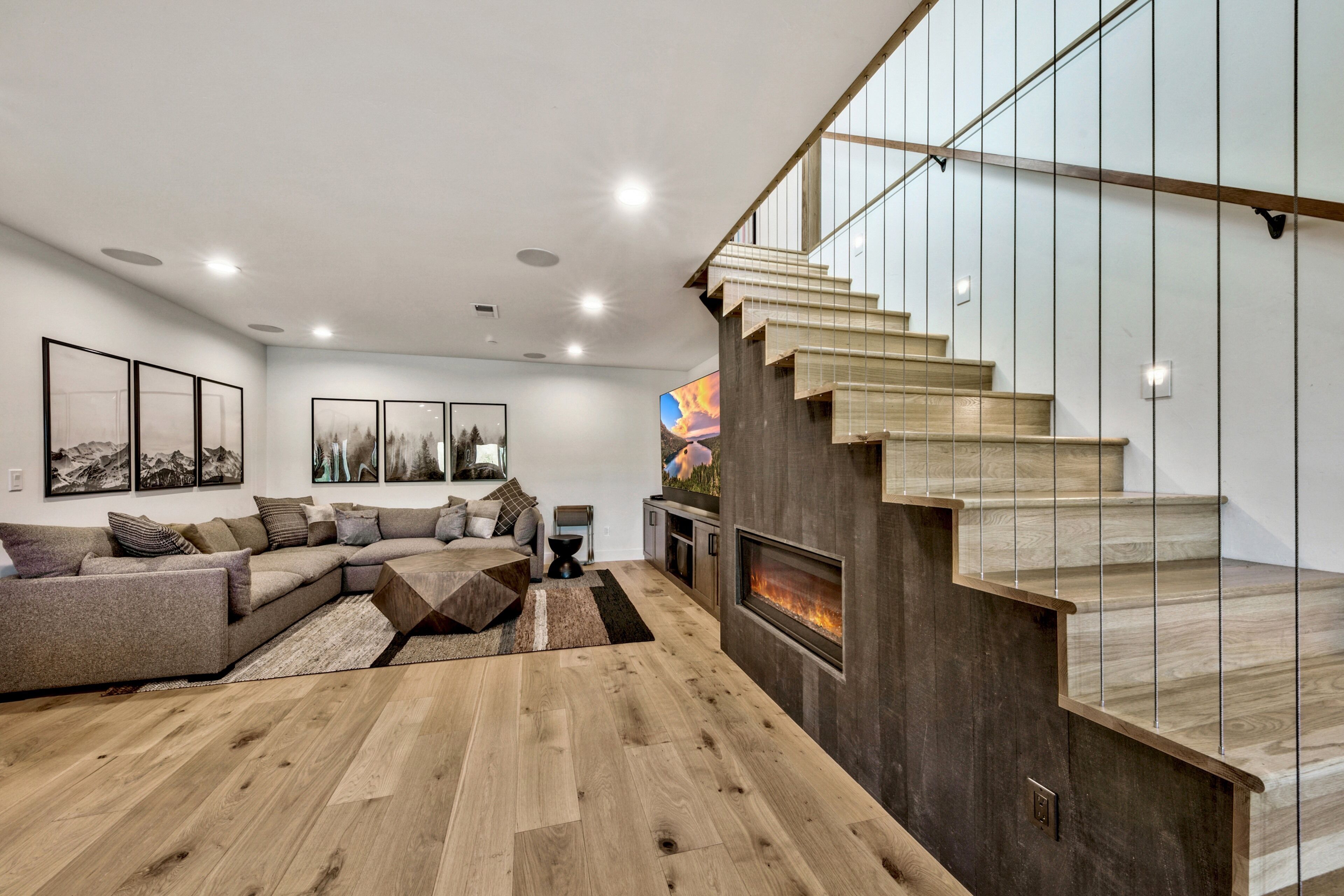 A modern living room with a wooden floor, a sectional sofa, a fireplace, and a staircase with glass railings.
