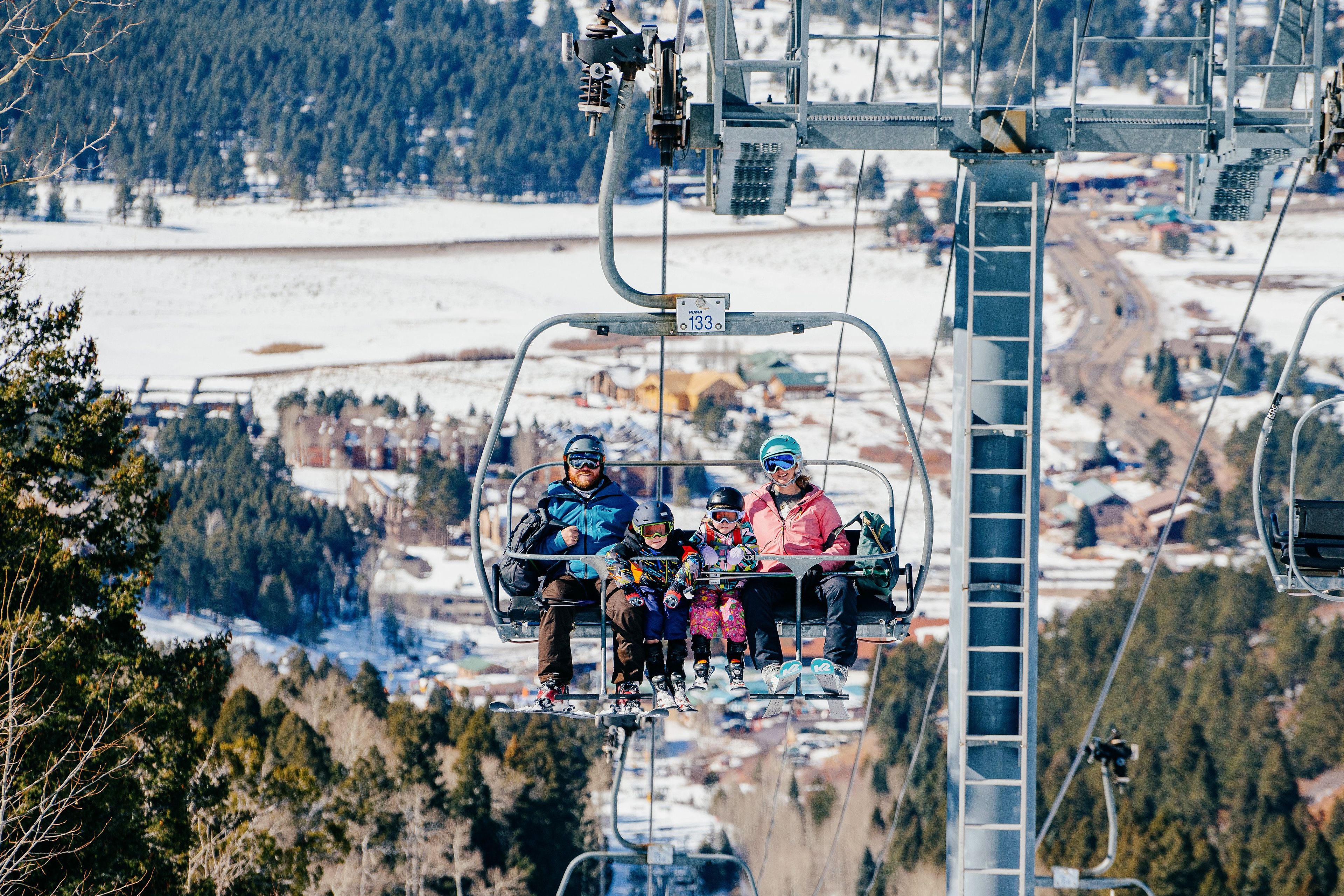 A ski lift with passengers, including a family with a child, against a backdrop of a snowy landscape and buildings.