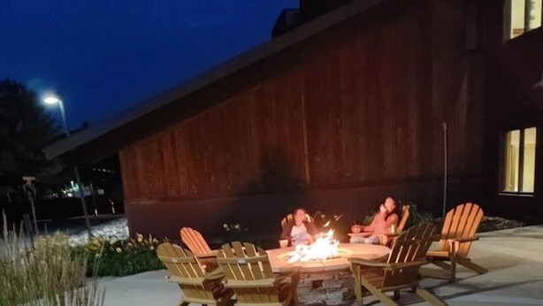 Un espacio al aire libre con sillas de madera y un brasero, rodeado de jardines cuidados y un edificio al fondo.