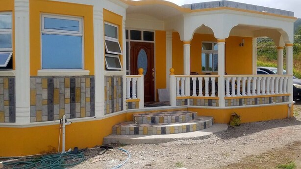 A two-story house with a yellow and white exterior, a front porch with a white balustrade, and a tiled entrance. - 🏝️ TAG Paradise Vacation Home | 1BR • A/C • Free Wi-Fi • Near Mero Beach (Roseau)