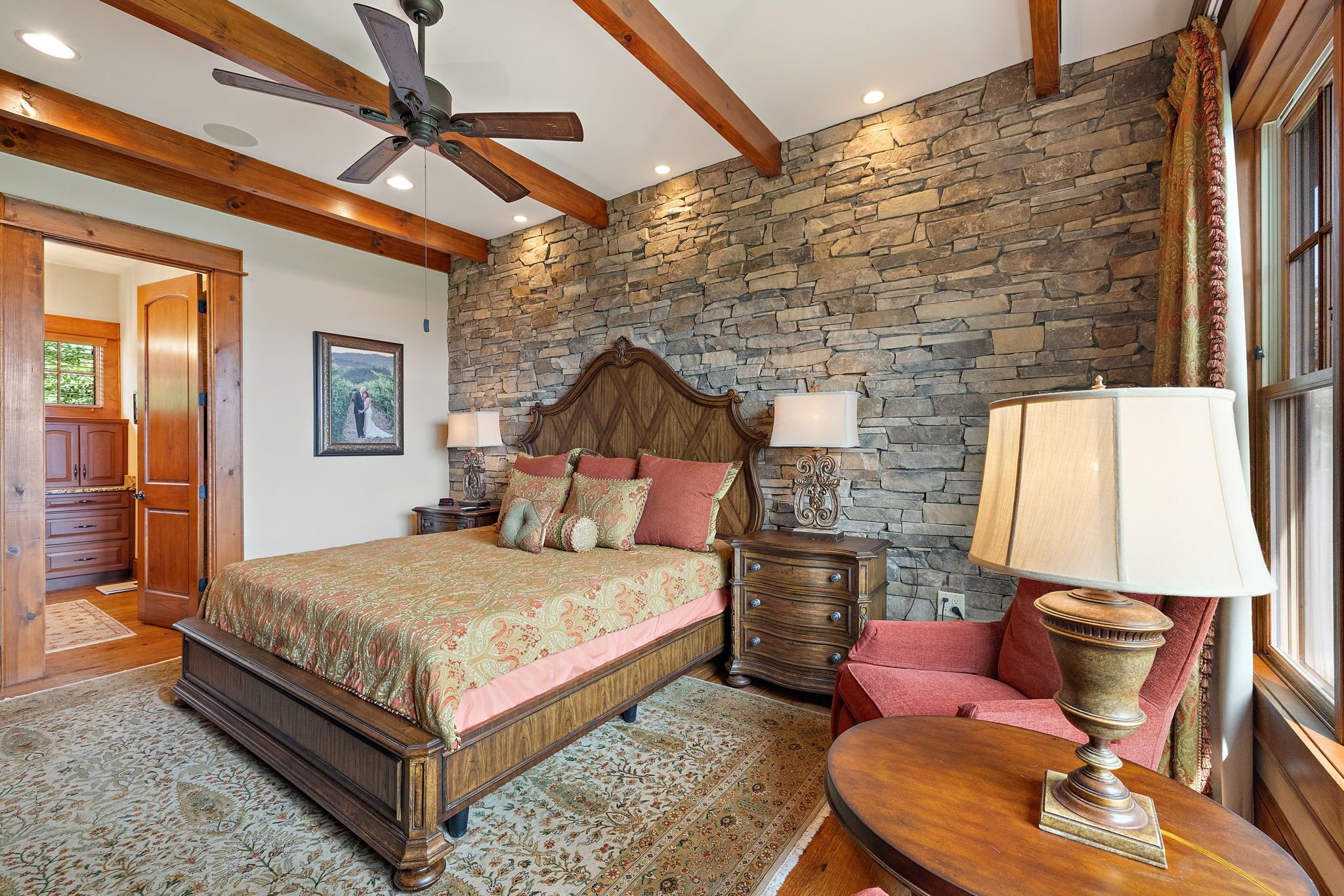 A bedroom with a stone wall, wooden ceiling beams, a bed with a patterned bedspread, two bedside tables with lamps, and a nightstand.