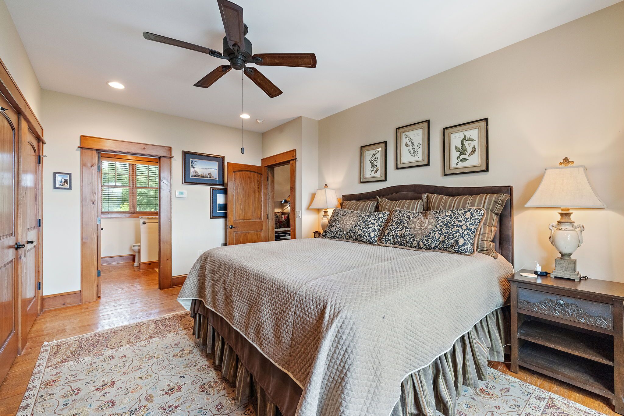 A bedroom with a large bed, a ceiling fan, wooden flooring, a door leading to a bathroom, and framed pictures on the wall.