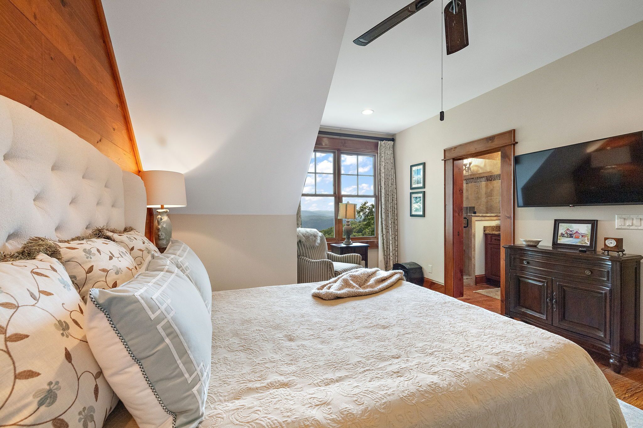 A bedroom with a large bed, a wooden headboard, a TV mounted on the wall, and a view of the outdoors through a window.