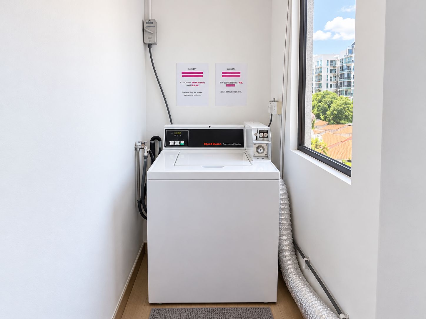 Una lavadora industrial compacta en una habitación pequeña con ventana, un enchufe eléctrico de pared y dos carteles informativos.