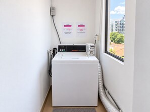 A compact commercial washer in a small room with a window, a wall-mounted electrical outlet, and two informational signs. - Aura Hotel Mackenzie (Singapore)