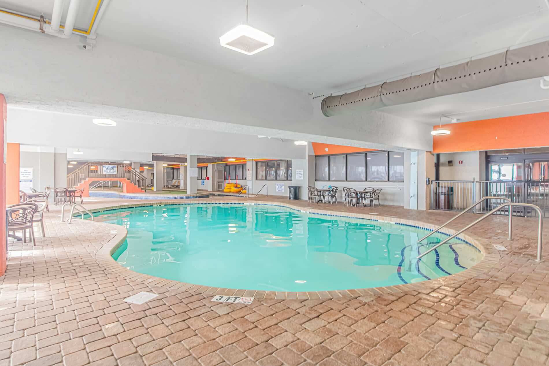 Una piscina interior con suelo de ladrillo, rodeada de zonas de estar y una escalera que conduce a niveles superiores.