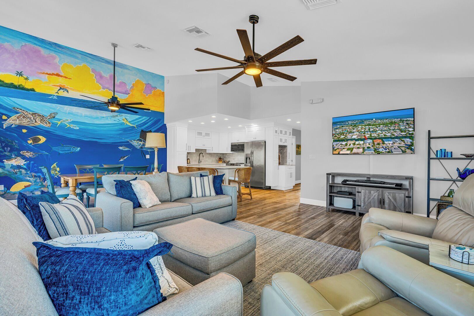 A modern living room with a large mural of a marine scene, a ceiling fan, and a view of the kitchen and dining area.