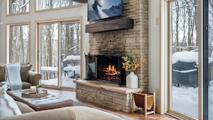 A cozy living room with a fireplace, a television mounted above it, and large windows offering a view of a snowy landscape.